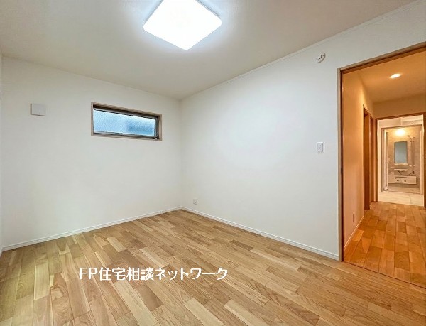 【洋室│6帖】  白い壁と温もりある木目調フローリングが、明るく清潔感のある空間を演出する洋室です。奥には洗面所などの水回りが配置されており、毎日の生活動線に配慮された使いやすい間取りです。 
